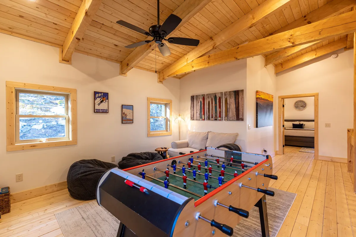 The photo features a foosball table at the center surrounded by plush seating and wooden beams, set beneath a sloped ceiling. Art on the walls and warm lighting create an intimate, cozy ambiance ideal for family gatherings and evening relaxation.