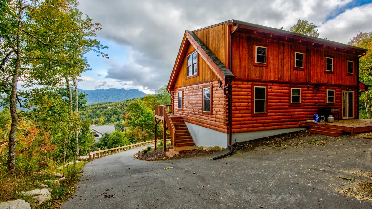 A classic log cabin structure is surrounded by lush greenery and mature trees. The driveway leads up to the house, with sweeping mountain views in the background. The deck and setting suggest year-round outdoor activities and family gatherings.