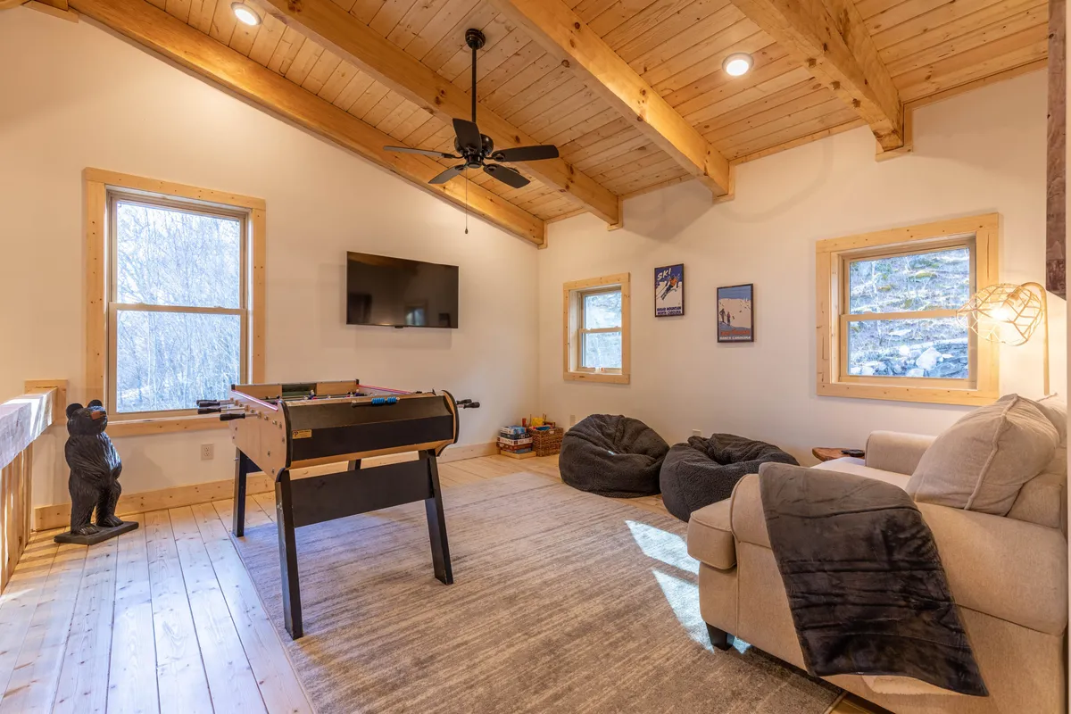 This image captures a lively loft area featuring a foosball table, comfortable seating, large windows that let in mountain light, and distinctive wooden beams. A decorative bear statue adds a playful touch, enhancing the cozy, family-friendly vibe. The scene conveys an inviting space where guests can gather, play games, or relax while enjoying beautiful views.