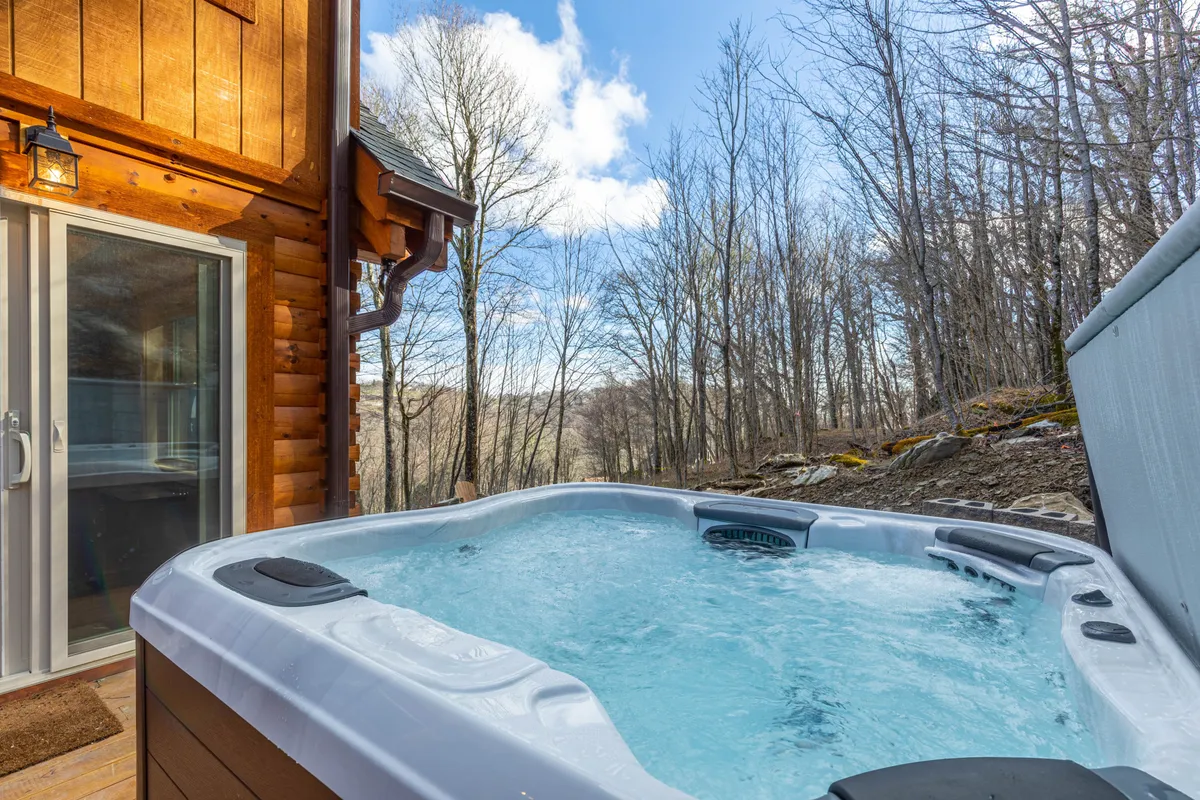 A serene exterior view featuring a wooden cabin with a prominent hot tub situated on the back deck, framed by lush natural scenery. The image captures guests enjoying the hot tub, with mountain views in the backdrop, highlighting a tranquil, inviting retreat.