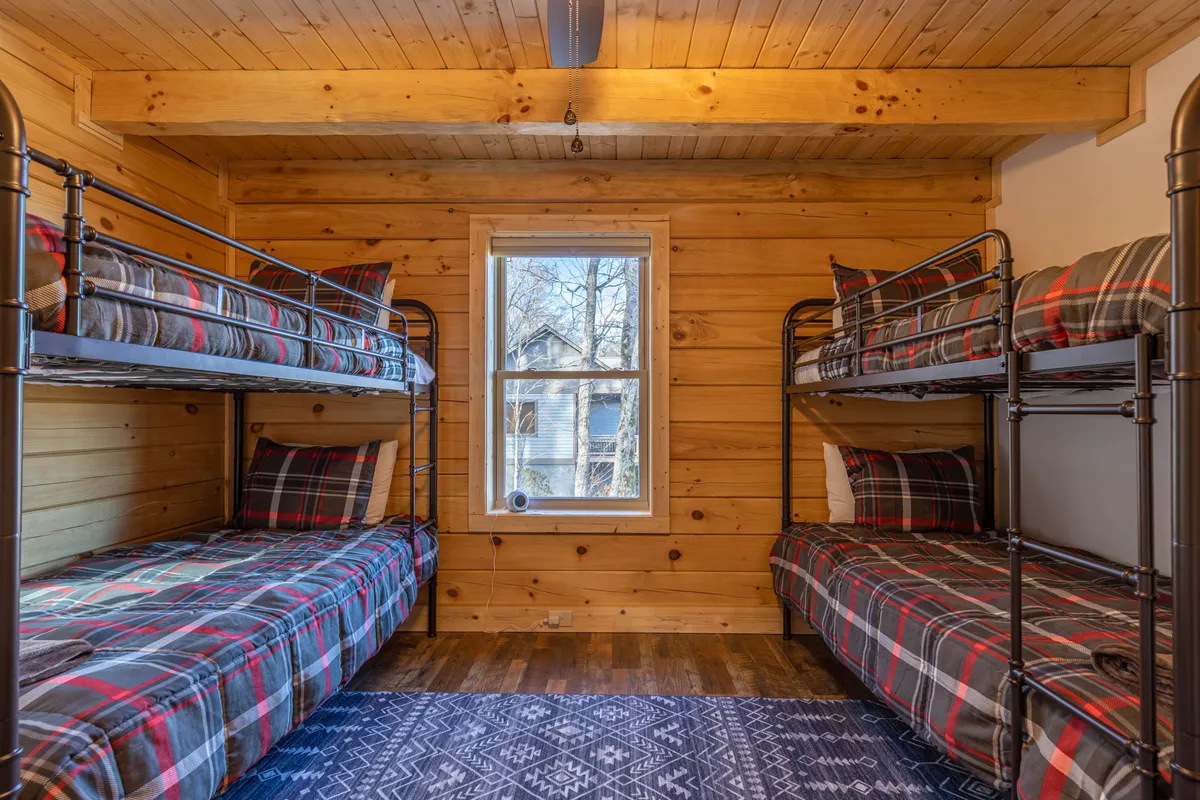 A cheerful bunk room features sturdy wooden bunk beds with plaid bedding, a window letting in natural light, and rustic wooden walls that create a cozy, playful atmosphere for kids.
