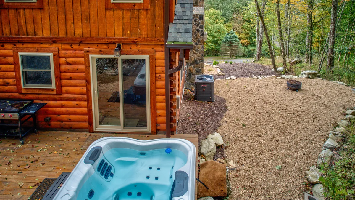 The image shows a wooden deck with a hot tub, a stone wall, and surrounding greenery. The tranquil setup invites relaxation, with space for outdoor dining or gathering around a fire pit, underlining the deck's versatility and scenic appeal.