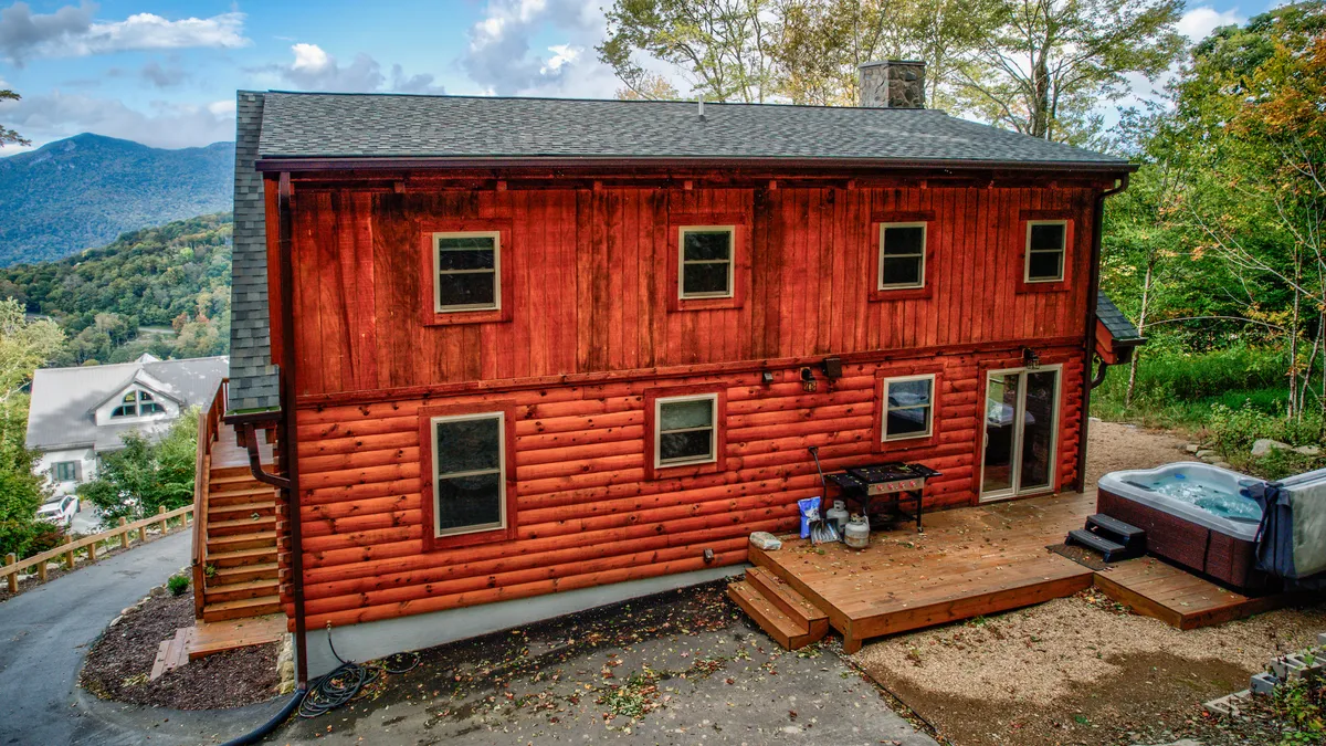 The image shows a cozy log cabin exterior with a hot tub on the wooden deck, surrounded by trees. The inviting setup is ideal for socializing, relaxing after exploring, or enjoying mountain air with friends and family.