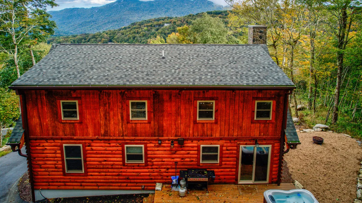 A log cabin exterior with a spacious deck overlooks sweeping mountain views. The inviting hot tub on the deck is the focal point, surrounded by comfortable seating, with the mountains in the distance hinting at outdoor adventure. The setting is serene, perfect for unwinding after a day of exploration.