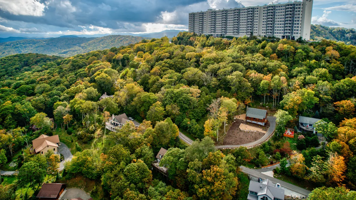 A wide shot featuring a tall building in the background, clusters of trees, a winding road, smaller homes, and an open area with a playground. The scene reflects a vibrant, active community set against a natural mountainous backdrop, suggesting outdoor adventure and nearby attractions.