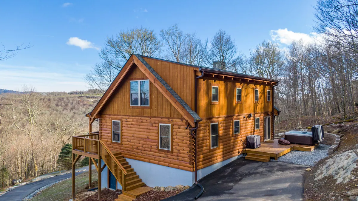 A cozy outdoor scene with a log cabin, wooden stairs leading to a spacious deck, and a hot tub nestled among the surrounding trees. The deck invites guests to relax in the hot tub and soak in views of the forest and sunsets, with hints of space for gatherings.