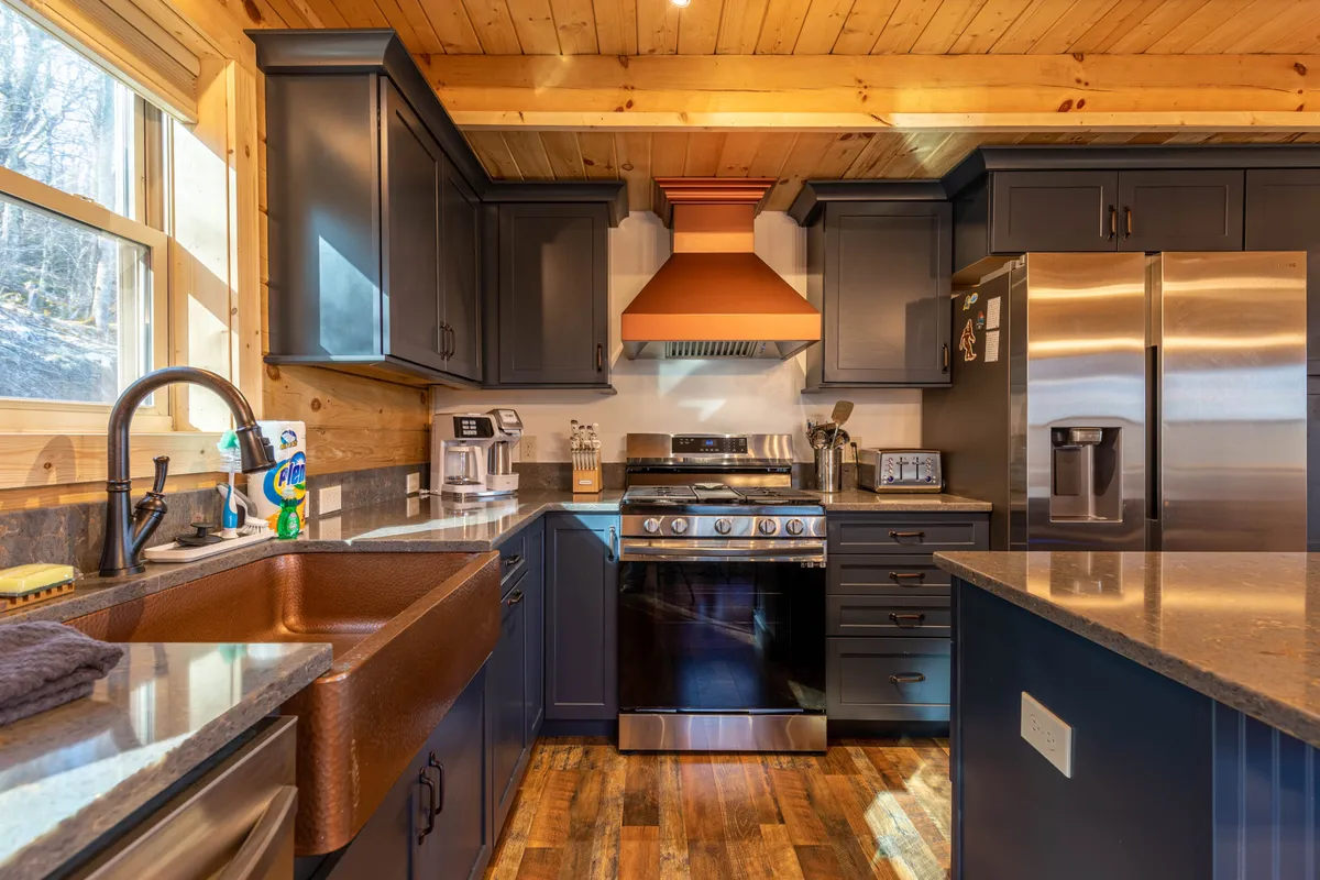 A closer look at the well-equipped kitchen featuring a striking copper farm sink, dark modern cabinetry, stainless steel appliances, granite countertops, and a rustic wooden ceiling. The space feels both luxurious and inviting, ideal for meal prep and entertaining.