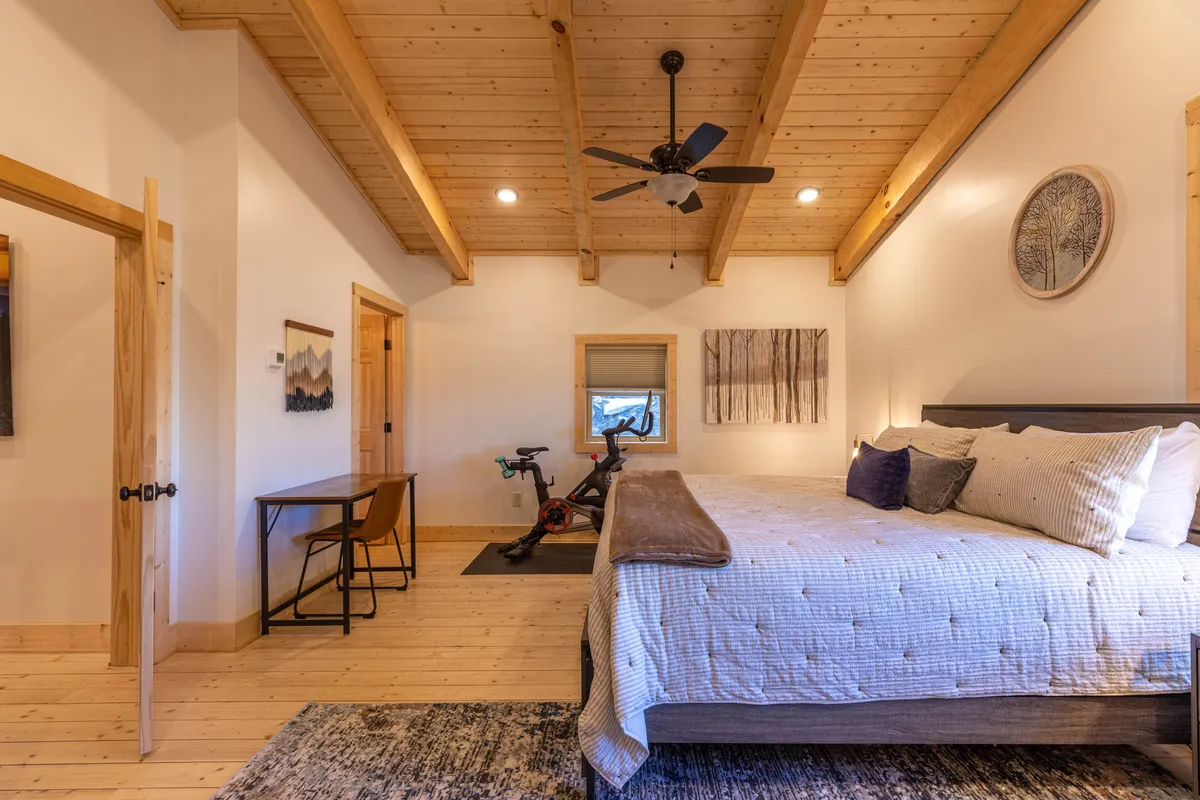 A spacious king bedroom featuring a comfortable bed, a desk and chair, and an exercise bike (Peloton). Well-lit by natural light, this tranquil retreat is tailored for both relaxation and fitness routines.