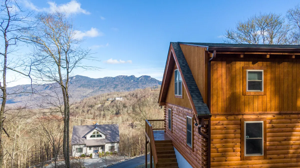 This exterior image captures two neighboring log cabin structures nestled in a mountain landscape with a clear view of both houses and the tree line between them. The scene highlights the proximity of the two homes, inviting guests to imagine booking both for a shared group experience.