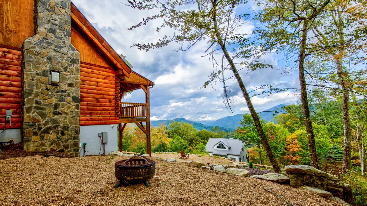 This image features a log cabin structure with a stone chimney nestled amidst vibrant autumn foliage and distant mountain views. A fire pit and porch set the scene for outdoor gatherings, with the landscape transitioning into a tapestry of fall colors.